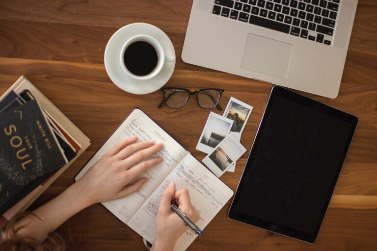 A woman writing on a notebook on top of a table with laptop, ipad, coffee, books, and photos on top