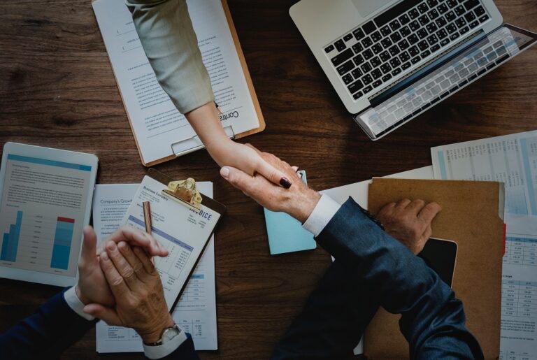 Two professionals shaking hands over a table with laptops, symbolizing collaboration and agreement in a business setting.