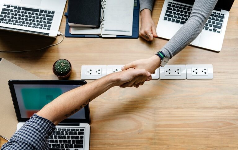 Two individuals shaking hands over a table, with laptops positioned on the surface, symbolizing collaboration and agreement.