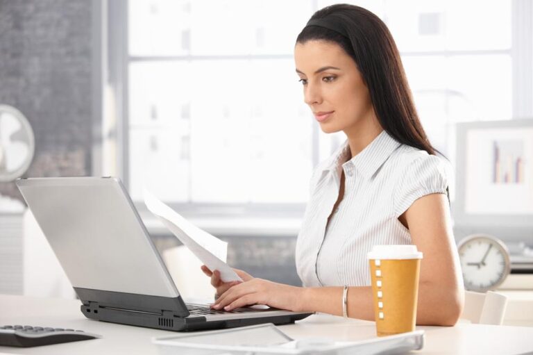 A woman at a desk, sipping coffee while working on her laptop, focused and engaged in her tasks.